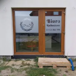 Wejście do biura rachunkowego z brązowymi drzwiami, oklejonymi szybami z logo firmy i napisem 'Biuro Rachunkowe' oraz numerem telefonu, widoczny drewniany podest i trawa.
