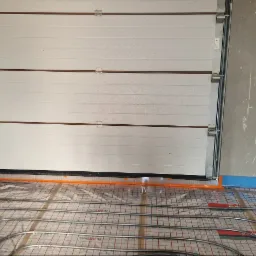 Montaż rur grzewczych na siatce w pomieszczeniu z bramą garażową, widoczne nieotynkowane ściany i wystające przewody elektryczne.