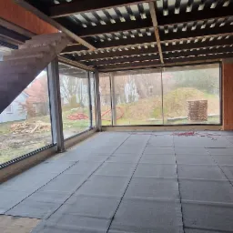 Przygotowanie podłoża pod wylewkę: rozłożone płyty izolacyjne na podłodze w surowym stanie, widoczne okna z widokiem na zewnątrz i konstrukcja stropu.