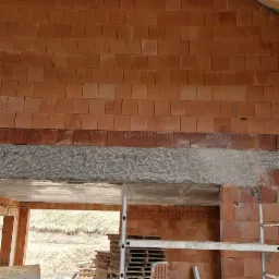 Wnętrze budynku w trakcie budowy, widoczna ściana z czerwonych cegieł, drewniana konstrukcja dachu i rusztowanie.