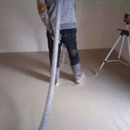 Aplikacja wylewki samopoziomującej w pomieszczeniu, widoczny pracownik w ubrudzonych spodniach i kaloszach, trzymający wąż, oraz laser krzyżowy na statywie, wyznaczający poziom wylewki.