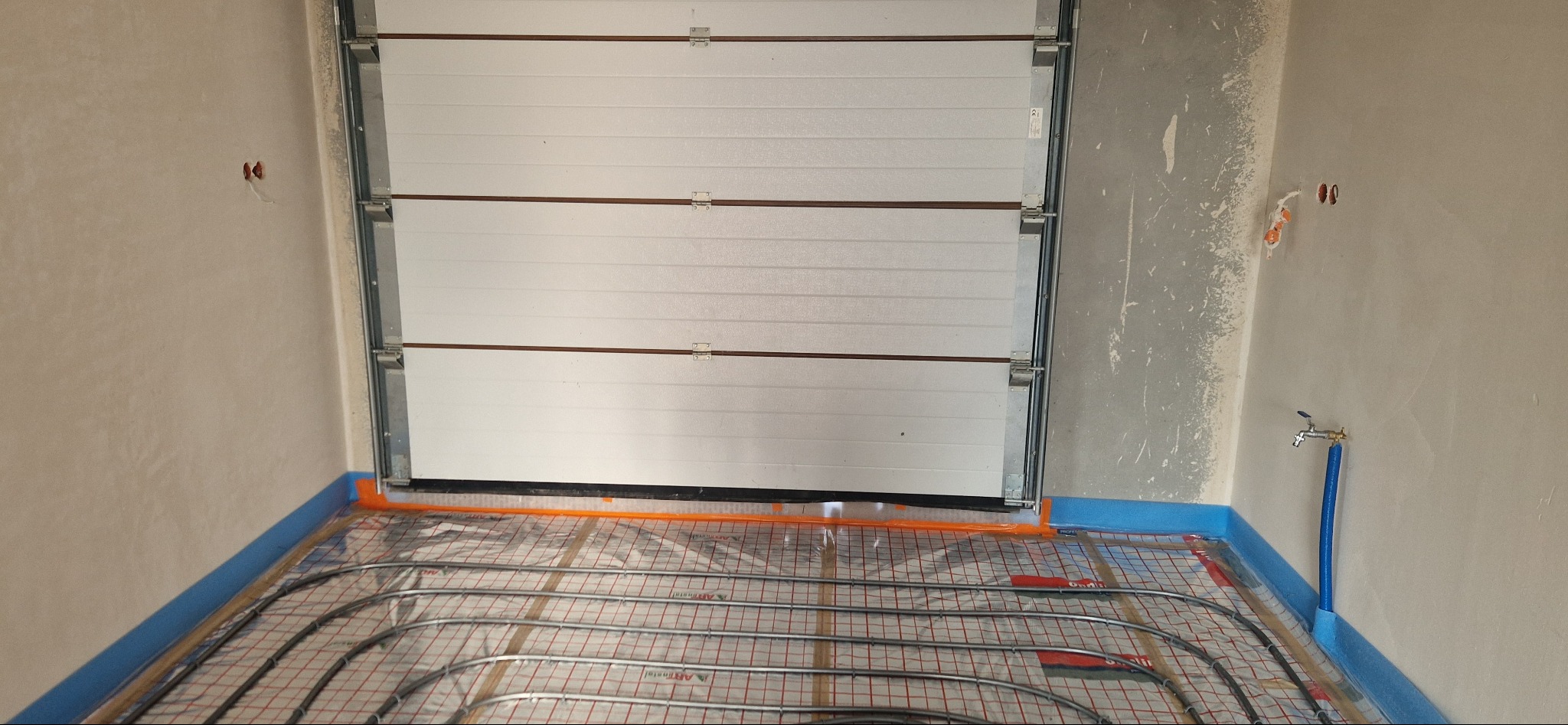 Montaż rur grzewczych na siatce w pomieszczeniu z bramą garażową, widoczne nieotynkowane ściany i wystające przewody elektryczne.
