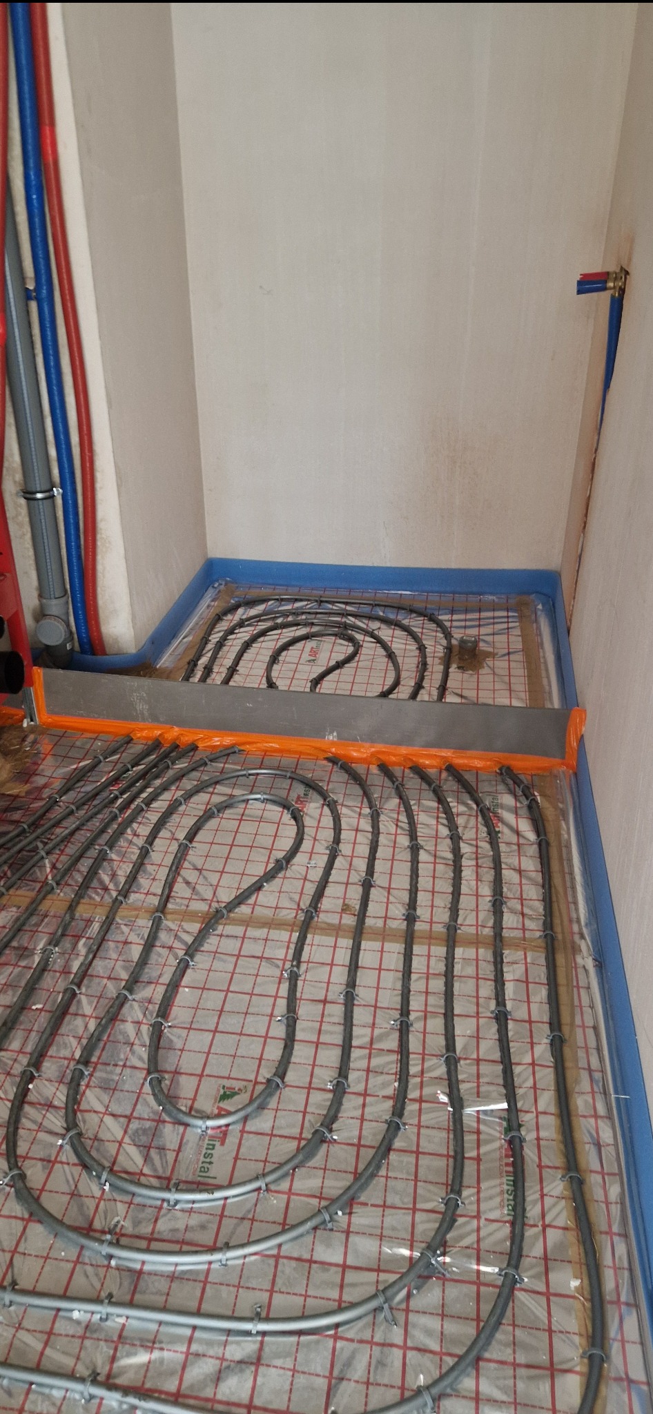 Rozłożone rury ogrzewania podłogowego na siatce z folią, przygotowane do zalania wylewką, widoczne mocowania rur i listwa dylatacyjna przy ścianie.