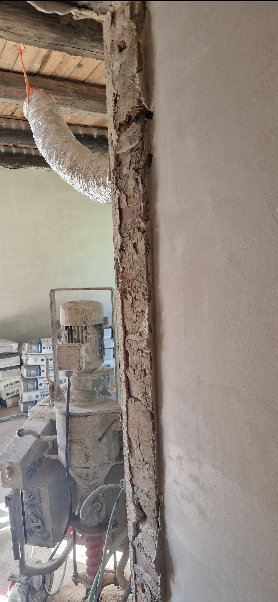 Przygotowanie ściany pod tynkowanie maszynowe: odsłonięta cegła na fragmencie ściany obok maszyny do tynkowania, widoczny przewód wentylacyjny i worki z zaprawą w tle.