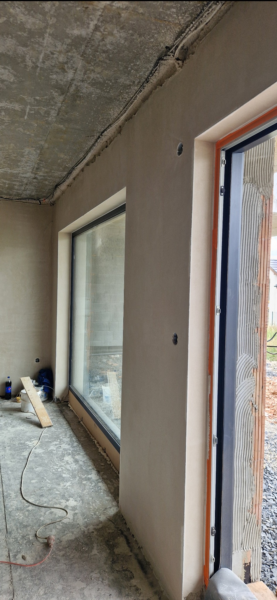 Wnętrze w stanie surowym z oknem i drzwiami, ściany pokryte świeżym tynkiem, widoczne niedoskonałości i nierówności, na podłodze resztki materiałów budowlanych i przewód elektryczny.