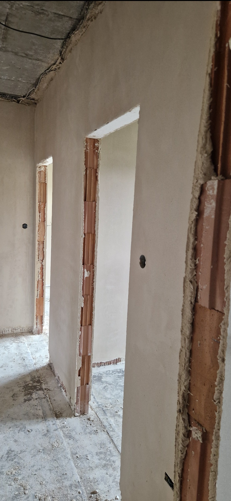 Surowe, otynkowane ściany wewnętrzne budynku w trakcie prac wykończeniowych, widoczne otwory drzwiowe z odsłoniętą cegłą i szary betonowy sufit z wystającymi kablami.