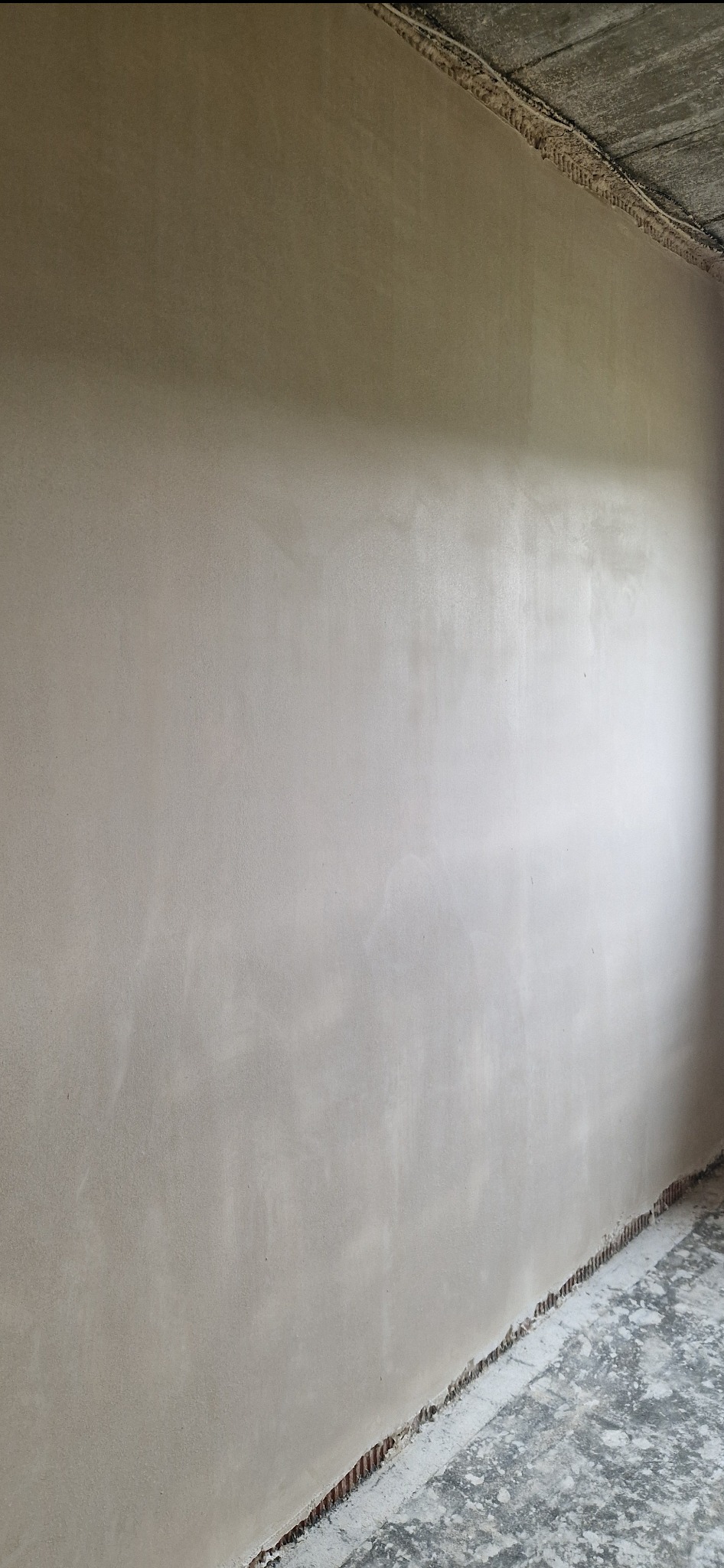 Gładka, jasna ściana po nałożeniu tynku maszynowego, widoczne niedoskonałości wykończenia przy podłodze i suficie, surowy betonowy strop.