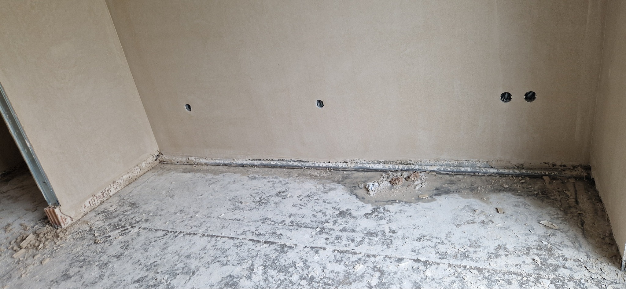 Pokój w trakcie remontu: świeżo otynkowane ściany w odcieniach beżu, widoczne puszki elektryczne w ścianie, surowa, nierówna posadzka z resztkami materiałów budowlanych.