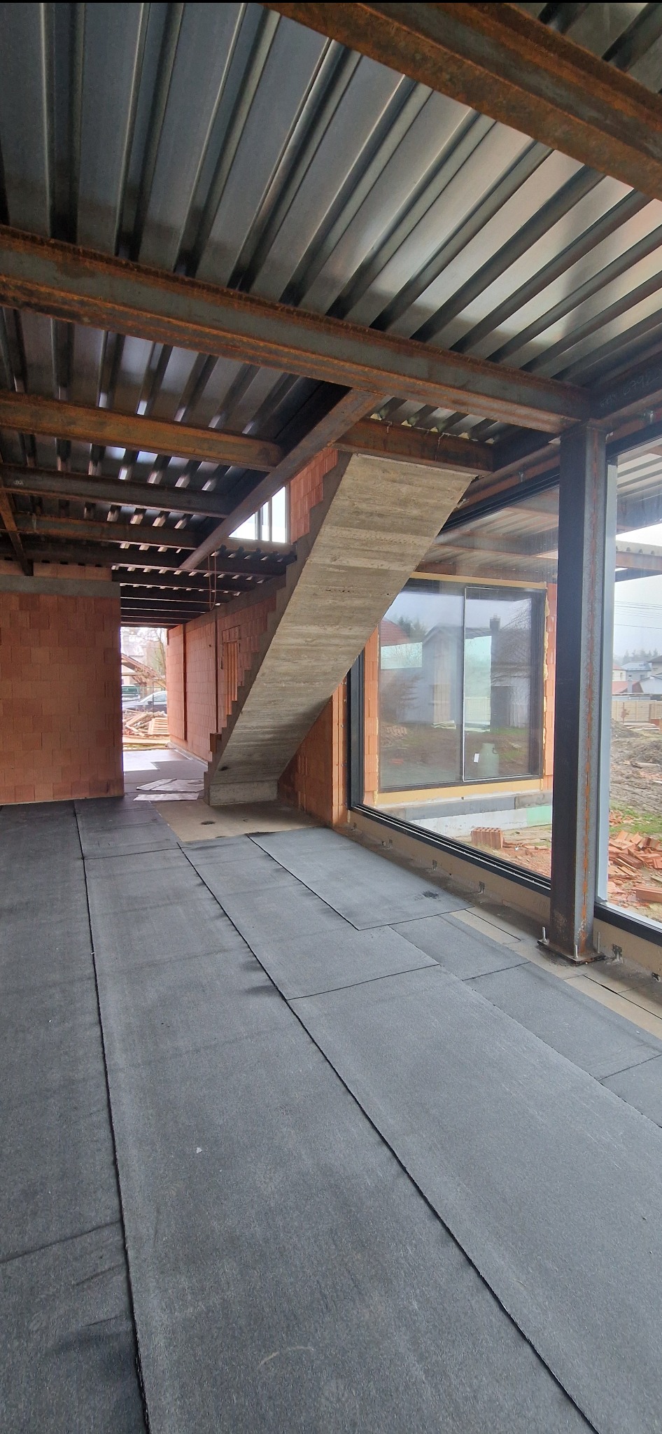 Wnętrze budynku w stanie surowym: podłoga pokryta czarną izolacją, betonowe schody bez balustrady, widoczne ceglane ściany i stalowa konstrukcja dachu.