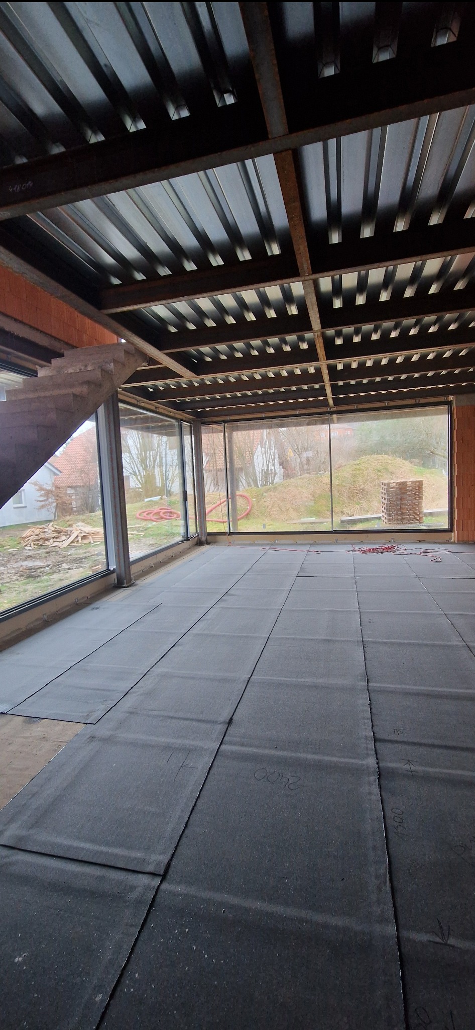 Przygotowanie podłoża pod wylewkę: rozłożone płyty izolacyjne na podłodze w surowym stanie, widoczne okna z widokiem na zewnątrz i konstrukcja stropu.