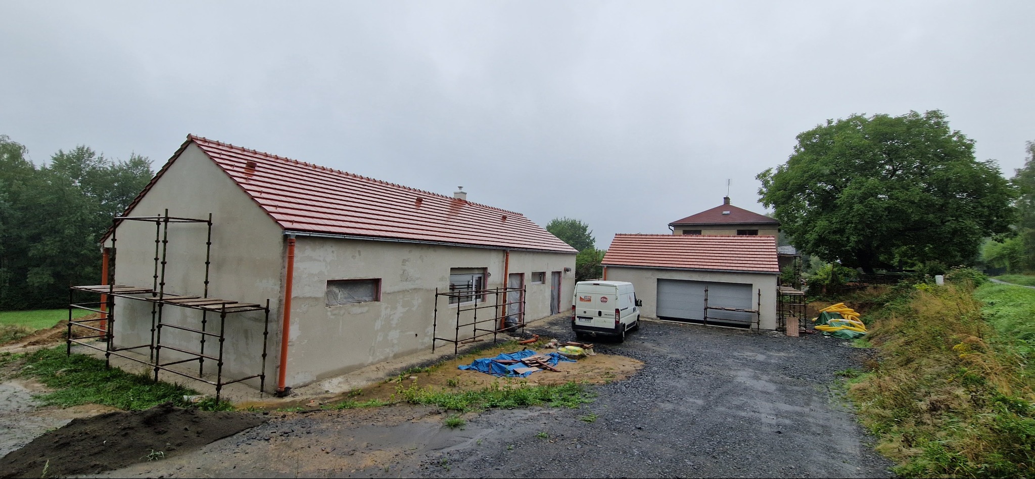 Budynek jednokondygnacyjny z nowym, czerwonym dachem z dachówki, częściowo otynkowany, z rusztowaniami przy ścianie, na podwórku zaparkowany biały bus.
