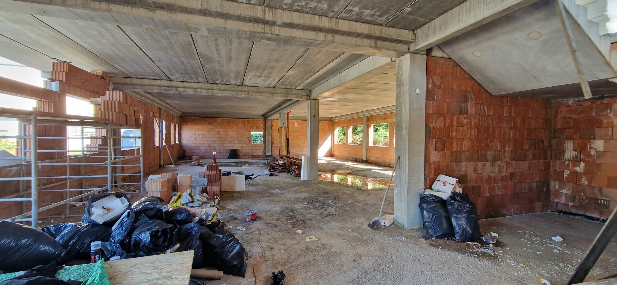 Surowy stan wnętrza budynku w trakcie budowy: mury z cegły, betonowy strop, prowizoryczne rusztowania, worki z odpadami budowlanymi i narzędzia.