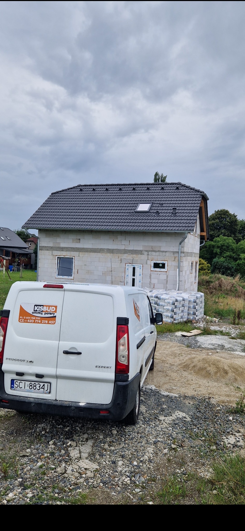 Surowy stan budynku z bloczków, z widocznym logo firmy Kisbud na zaparkowanym białym samochodzie dostawczym Peugeot Expert, obok sterta worków z materiałem budowlanym.
