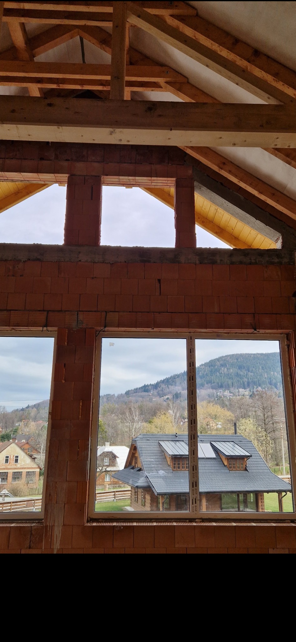 Surowy stan budynku w trakcie budowy: widok z wnętrza przez okna na krajobraz z domami i wzgórzami, widoczna konstrukcja więźby dachowej i murowane ściany z pustaków.