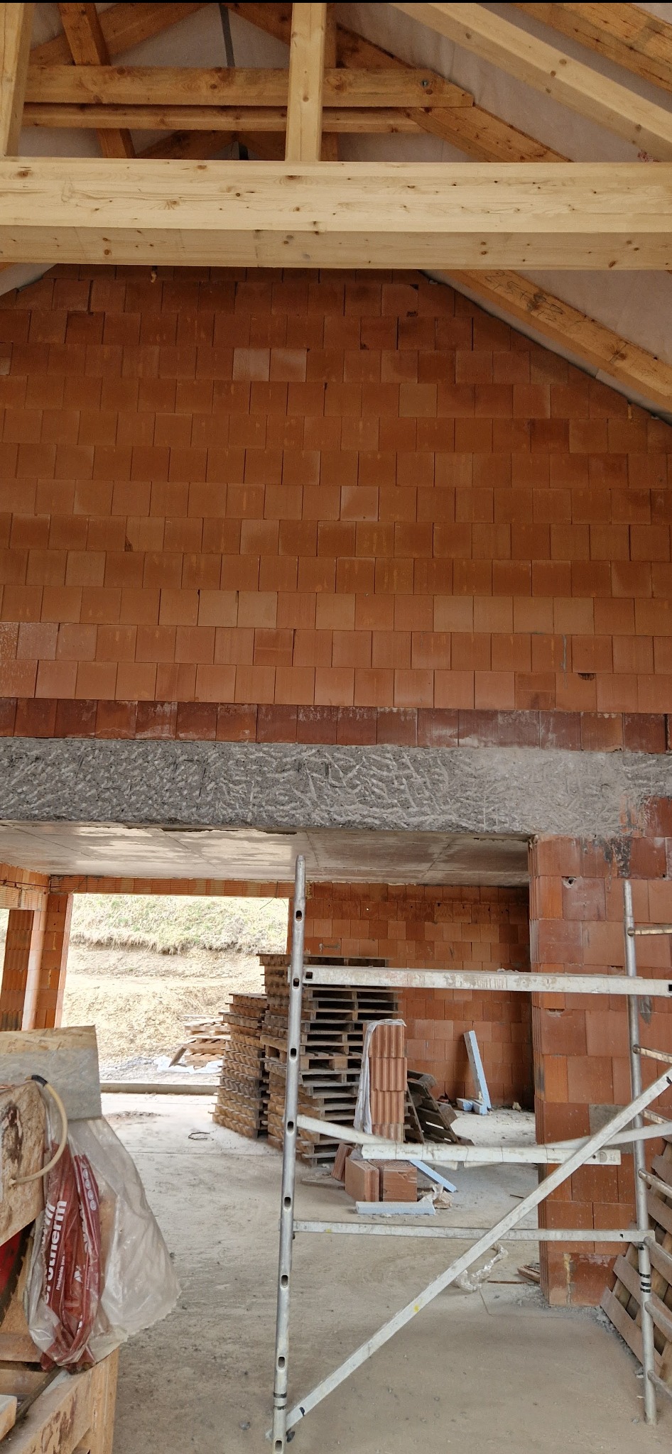 Wnętrze budynku w trakcie budowy, widoczna ściana z czerwonych cegieł, drewniana konstrukcja dachu i rusztowanie.