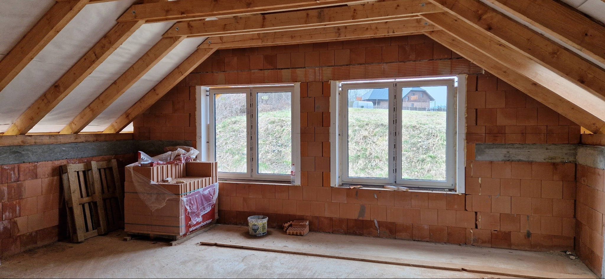 Poddasze w trakcie budowy: widoczne ceglane ściany, drewniana konstrukcja dachu z izolacją, dwa okna z widokiem na zewnątrz, paleta i cegły Wienerberger czekające na użycie.