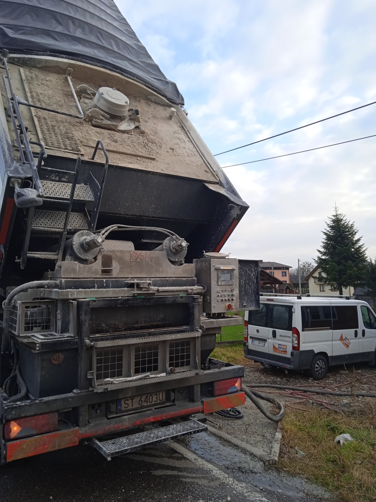 Ciężki sprzęt do wylewania posadzek, z widocznym podajnikiem i wężami, zaparkowany obok białego busa firmy budowlanej na tle domów i drzew.