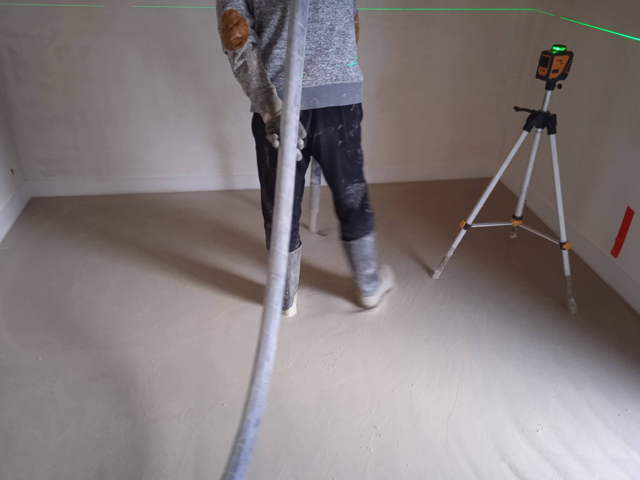 Aplikacja wylewki samopoziomującej w pomieszczeniu, widoczny pracownik w ubrudzonych spodniach i kaloszach, trzymający wąż, oraz laser krzyżowy na statywie, wyznaczający poziom wylewki.
