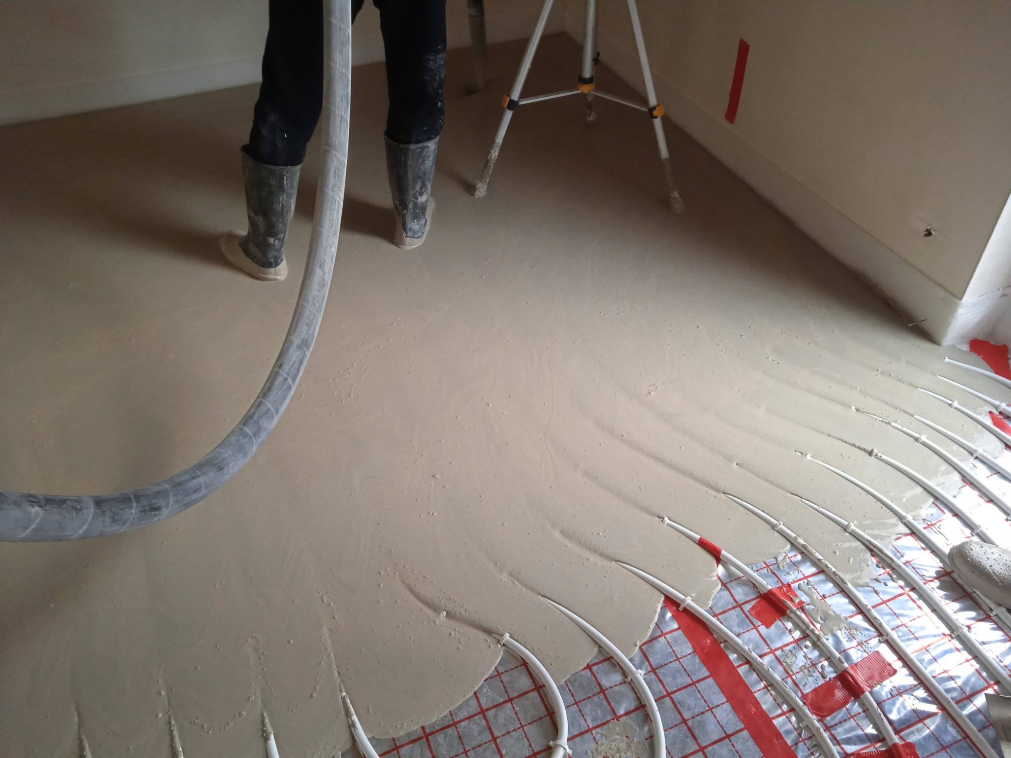 Wylewka podłogowa w trakcie rozprowadzania, widoczne rury ogrzewania podłogowego na folii z czerwoną kratą, osoba w gumowych butach z wężem do wylewki, statyw w tle.