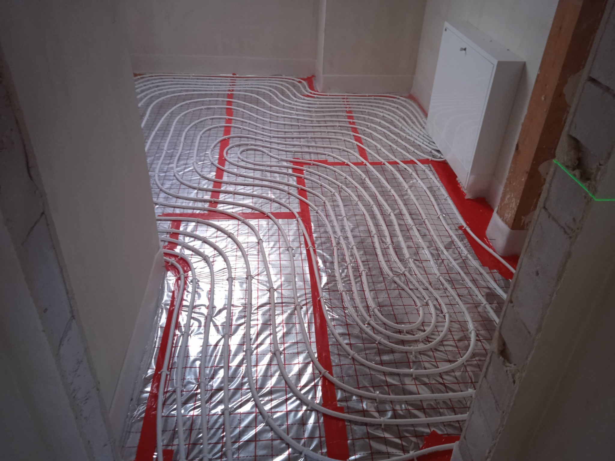 Białe rury ogrzewania podłogowego ułożone spiralnie na folii izolacyjnej z czerwoną kratką, widok z góry na surowy stan wykończenia wnętrza.