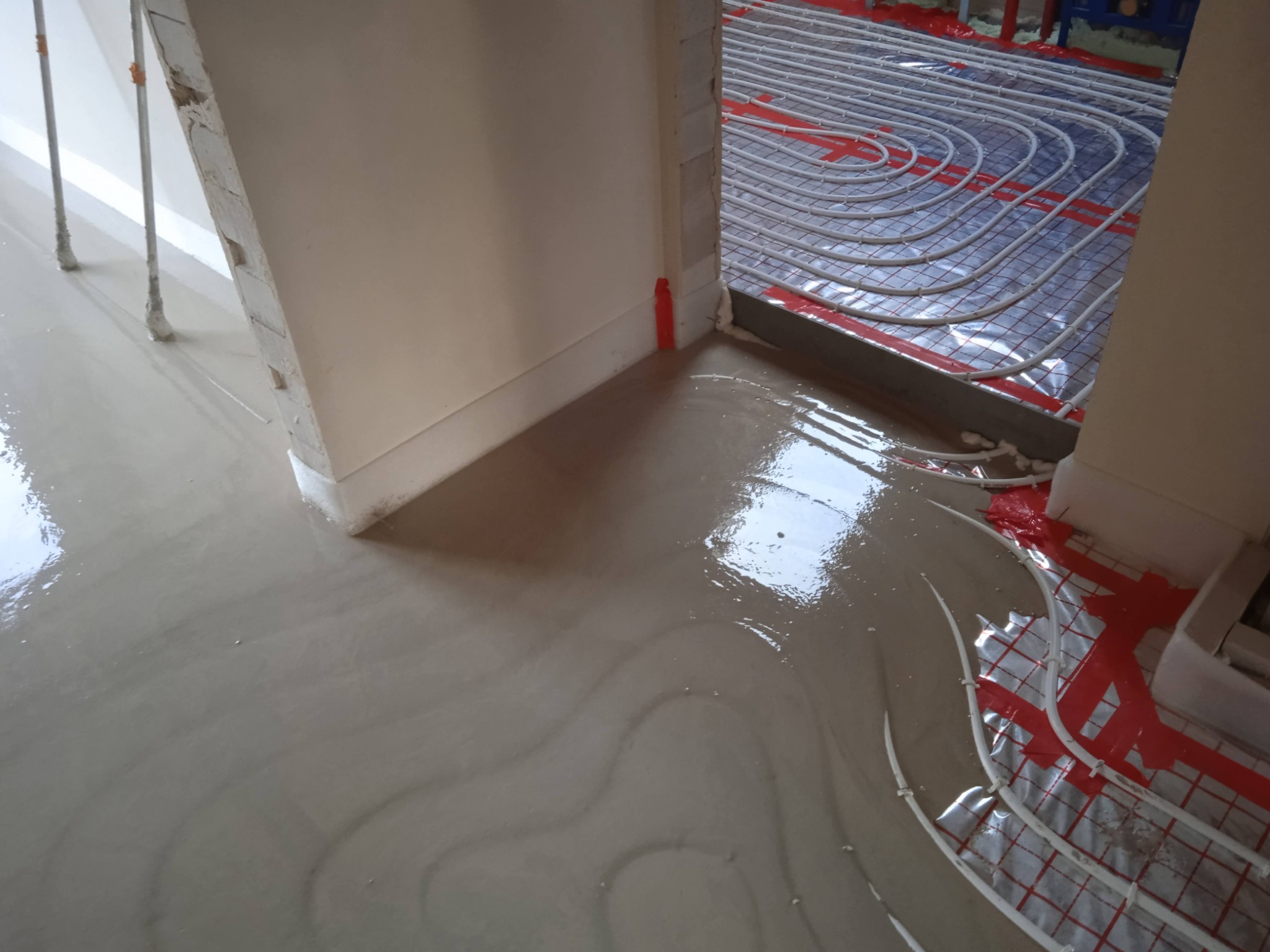 Świeżo wylana, błyszcząca posadzka samopoziomująca z widocznym systemem rur ogrzewania podłogowego na czerwonej folii, częściowo już zalanych, w trakcie prac wykończeniowych.