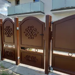Brązowa, metalowa brama wjazdowa z dekoracyjnymi, ażurowymi elementami, częściowo otwarta, z widoczną balustradą szklaną na balkonie budynku w tle.