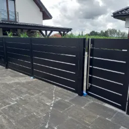 Nowoczesna, czarna brama wjazdowa z poziomymi, srebrnymi akcentami, zamontowana na posesji z kostki brukowej, widok na dom w tle pod pochmurnym niebem.