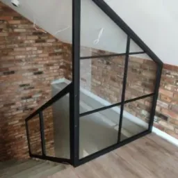 Szklana balustrada z czarnymi profilami, zamontowana na schodach w domu z ceglaną ścianą i drewnianą podłogą.