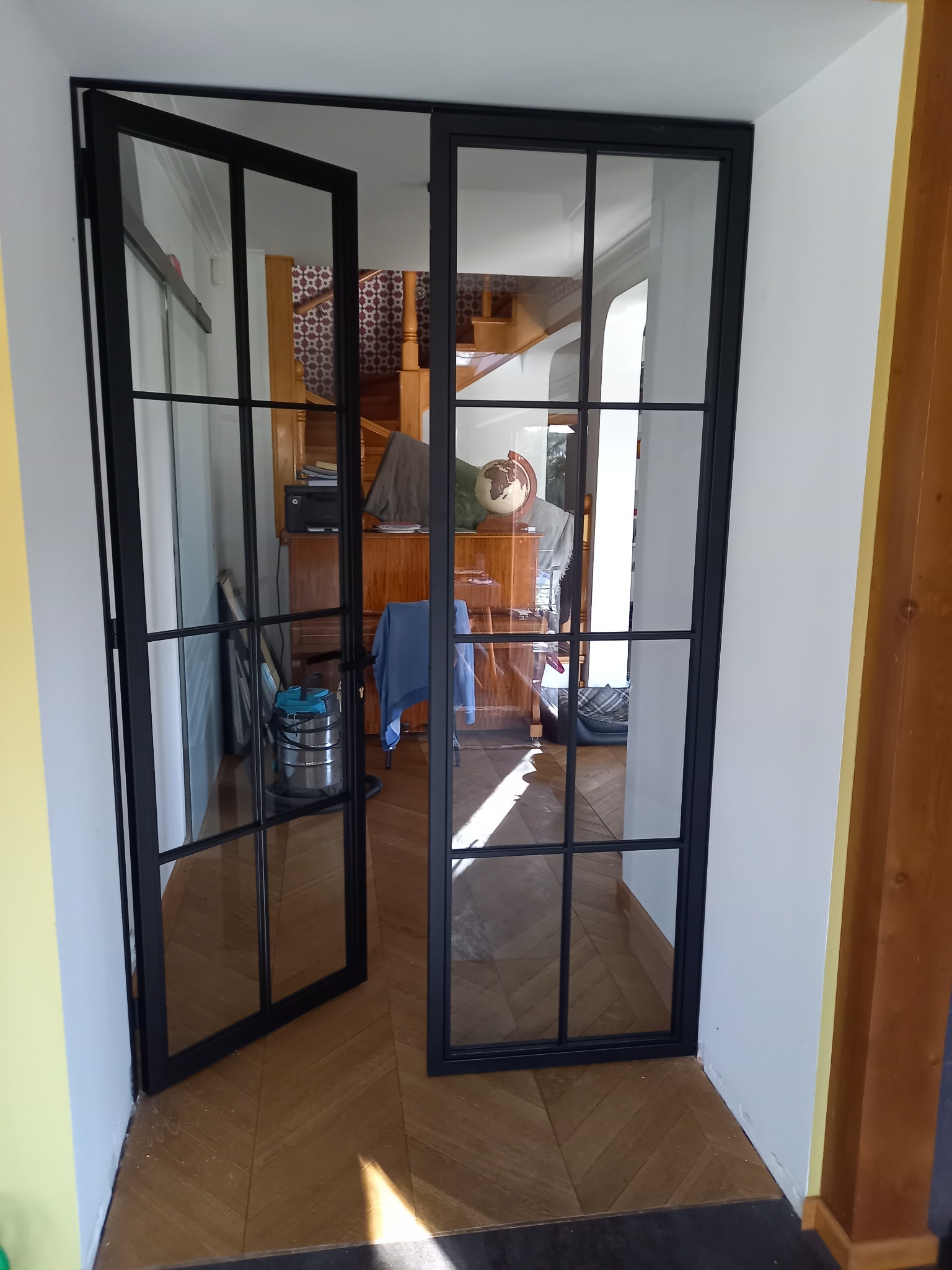 Nowoczesne, czarne, przeszklone drzwi wewnętrzne z podziałami szprosowymi, otwarte do wnętrza z parkietem ułożonym w jodełkę.