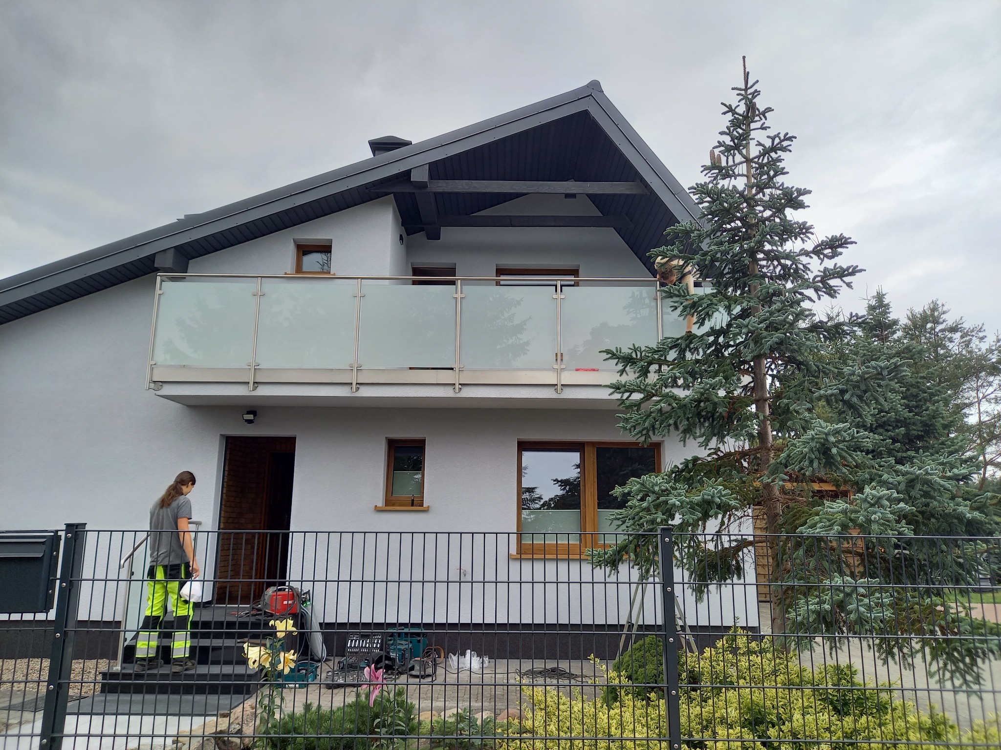 Dom w Leśniewie z balkonem zabezpieczonym balustradą ze szkła i stali nierdzewnej; monter w odblaskowych spodniach schodzi po schodach z narzędziami.