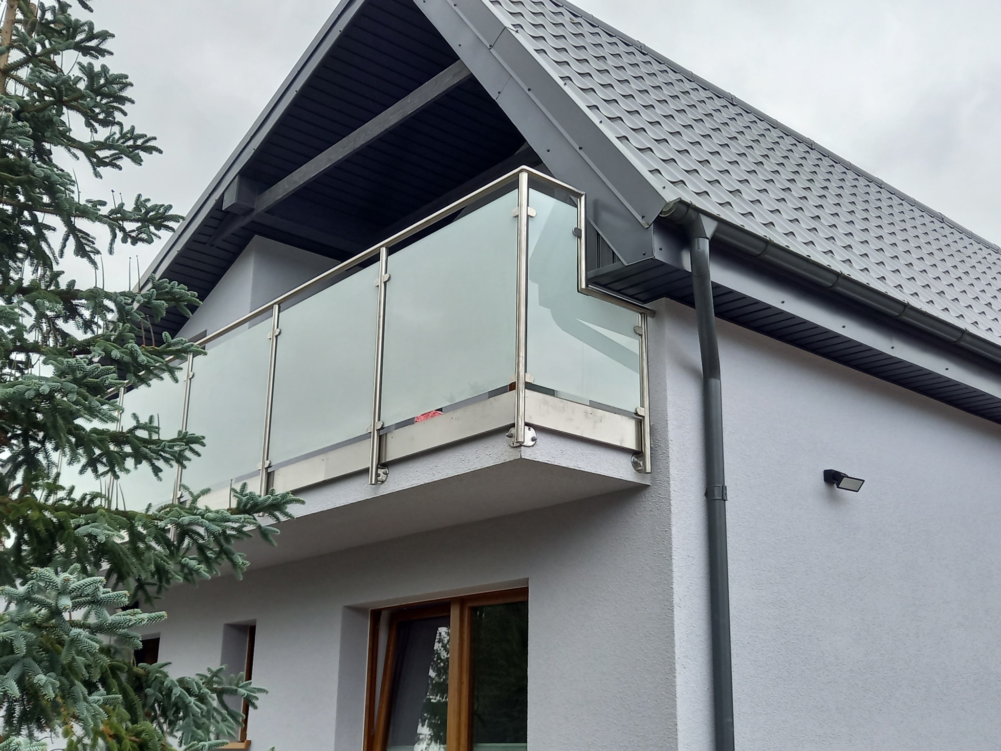 Nowoczesny balkon z przeszkloną balustradą i stalowymi słupkami, widok z dołu na tle szarego dachu i elewacji domu z oknami oraz zielonego drzewa iglastego.