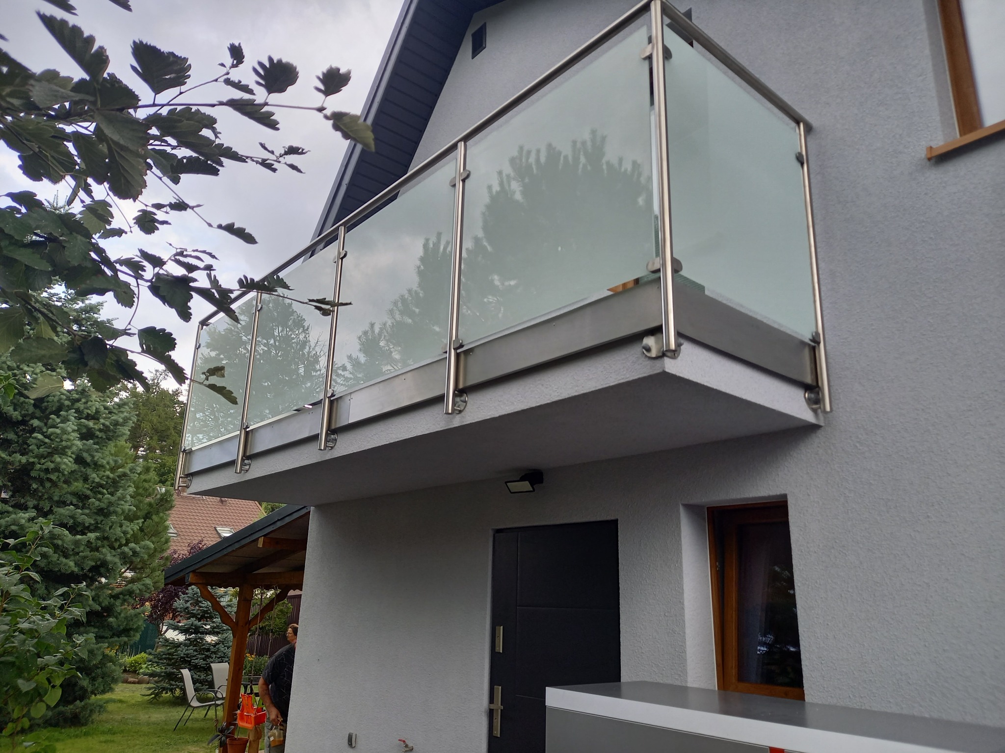 Nowoczesny balkon z szklaną balustradą i elementami ze stali nierdzewnej, widok z dołu na tle szarej elewacji domu i zieleni ogrodu.