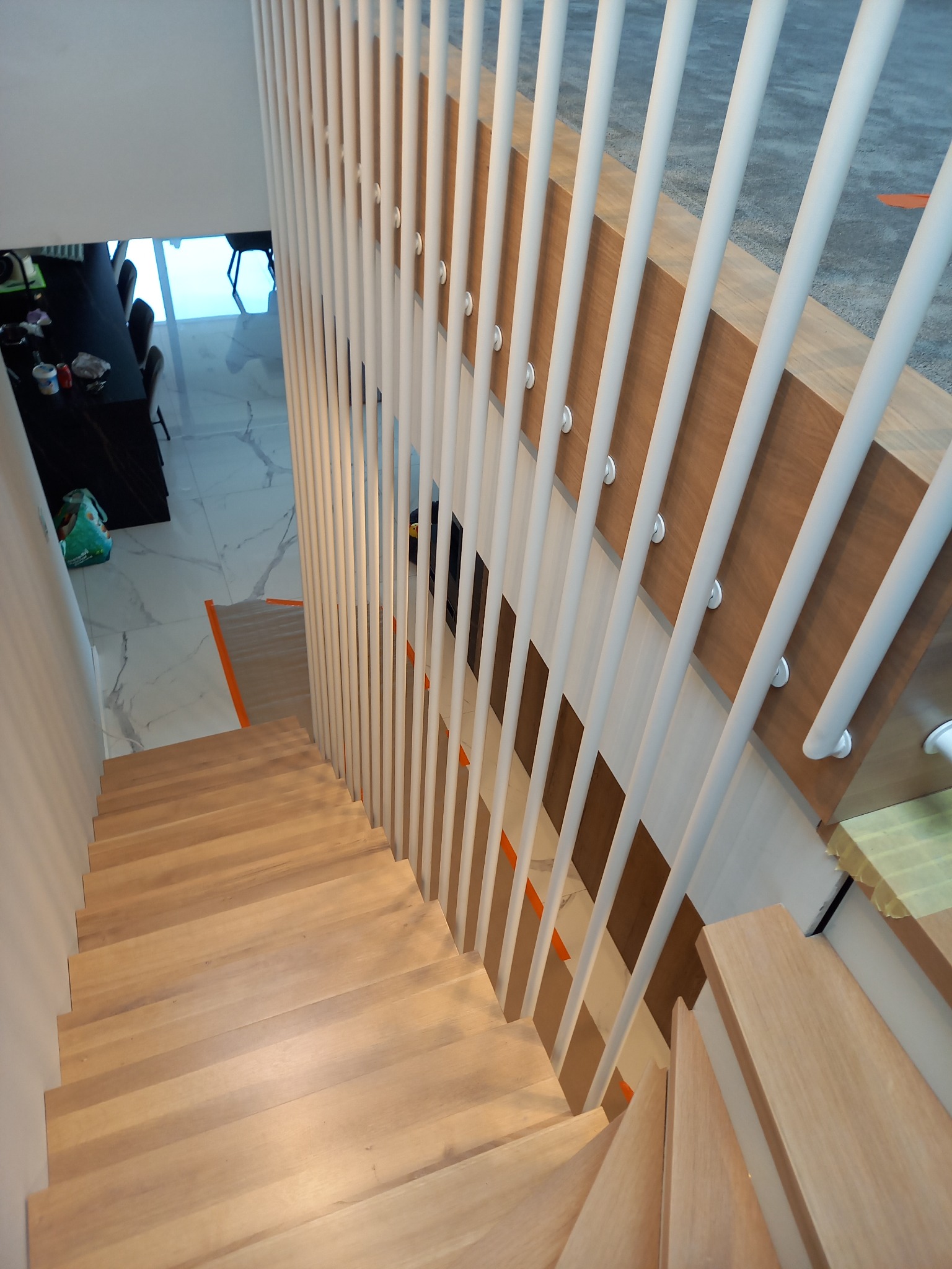 Nowoczesna drewniana klatka schodowa z białą poręczą z pionowych listew, widok z góry na białą marmurową podłogę i stół w tle.