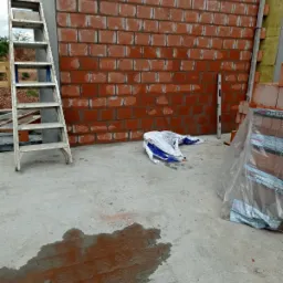 Wznoszenie ściany z czerwonej cegły, widoczna drabina aluminiowa, poziomica pionowa przy ścianie, paczka cegieł w folii i mokra plama na betonowej posadzce.