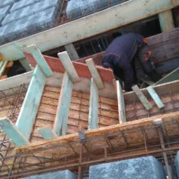 Prace zbrojarskie i szalunkowe pod fundamenty w Nowym Mieście nad Wartą, widoczne zbrojenie i drewniany szalunek, pracownik w kombinezonie wewnątrz konstrukcji.