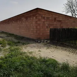 Surowa, wysoka ściana budynku w trakcie budowy z czerwonej cegły, widoczna z zewnątrz na tle piaszczystego terenu z porastającą trawą i drewnianym ogrodzeniem.