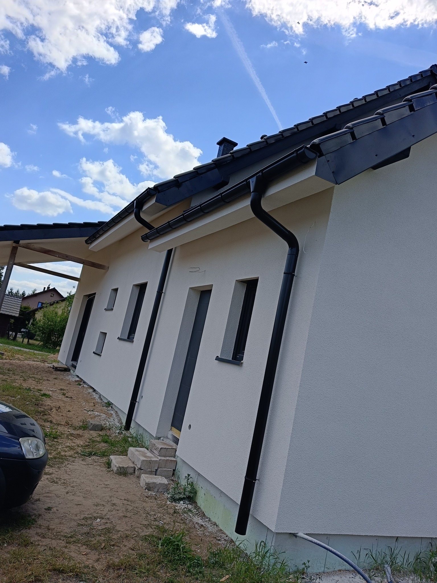Elewacja domu jednorodzinnego z czarnymi rynnami na tle błękitnego nieba. Widoczne okna i fragment dachu. Nierówny teren wokół budynku.