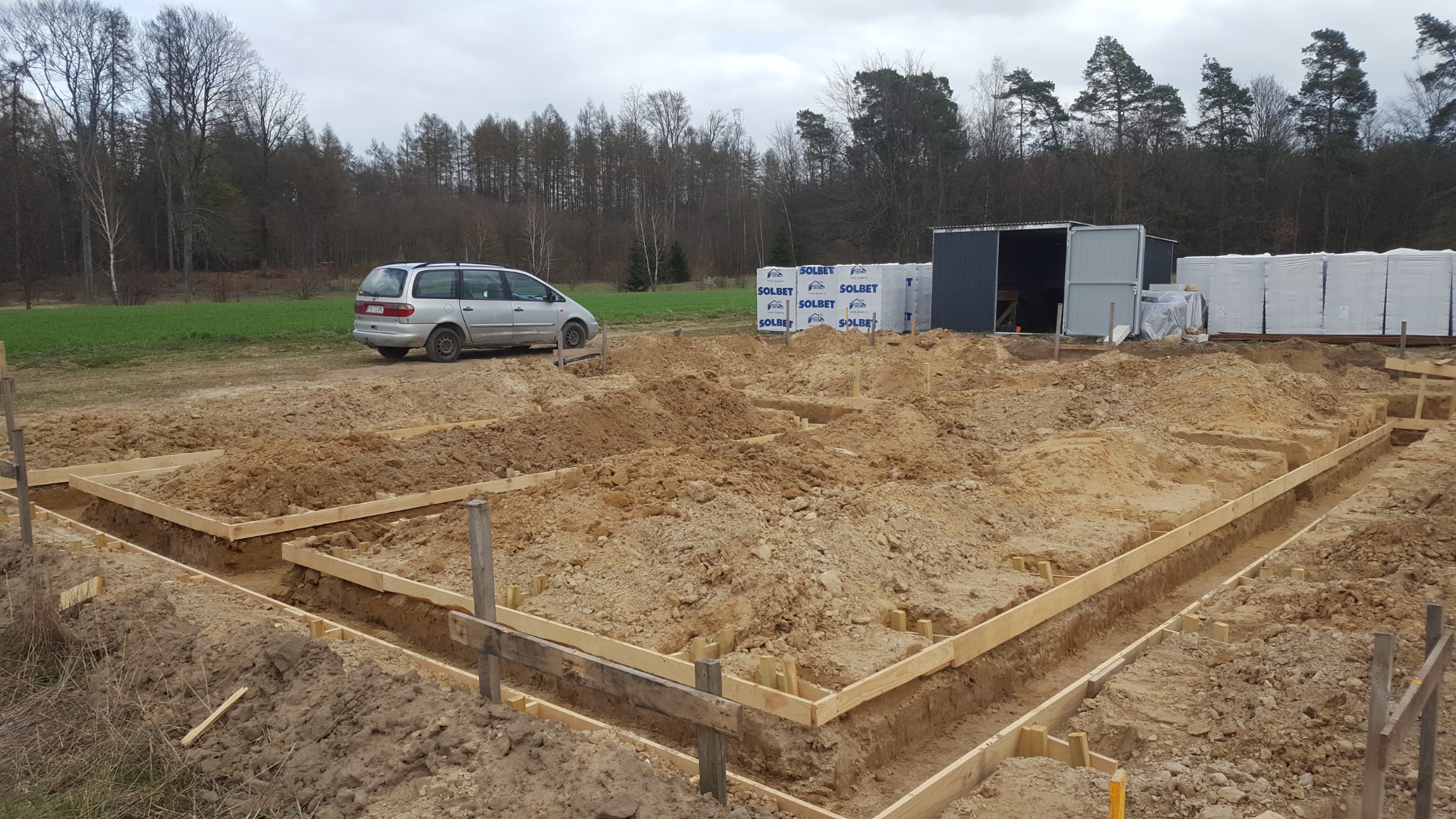 Przygotowanie fundamentów pod budowę domu: wykopane rowy zalane ziemią, obramowane deskami, widoczne bloczki SOLBET i zaparkowany srebrny samochód kombi w tle.
