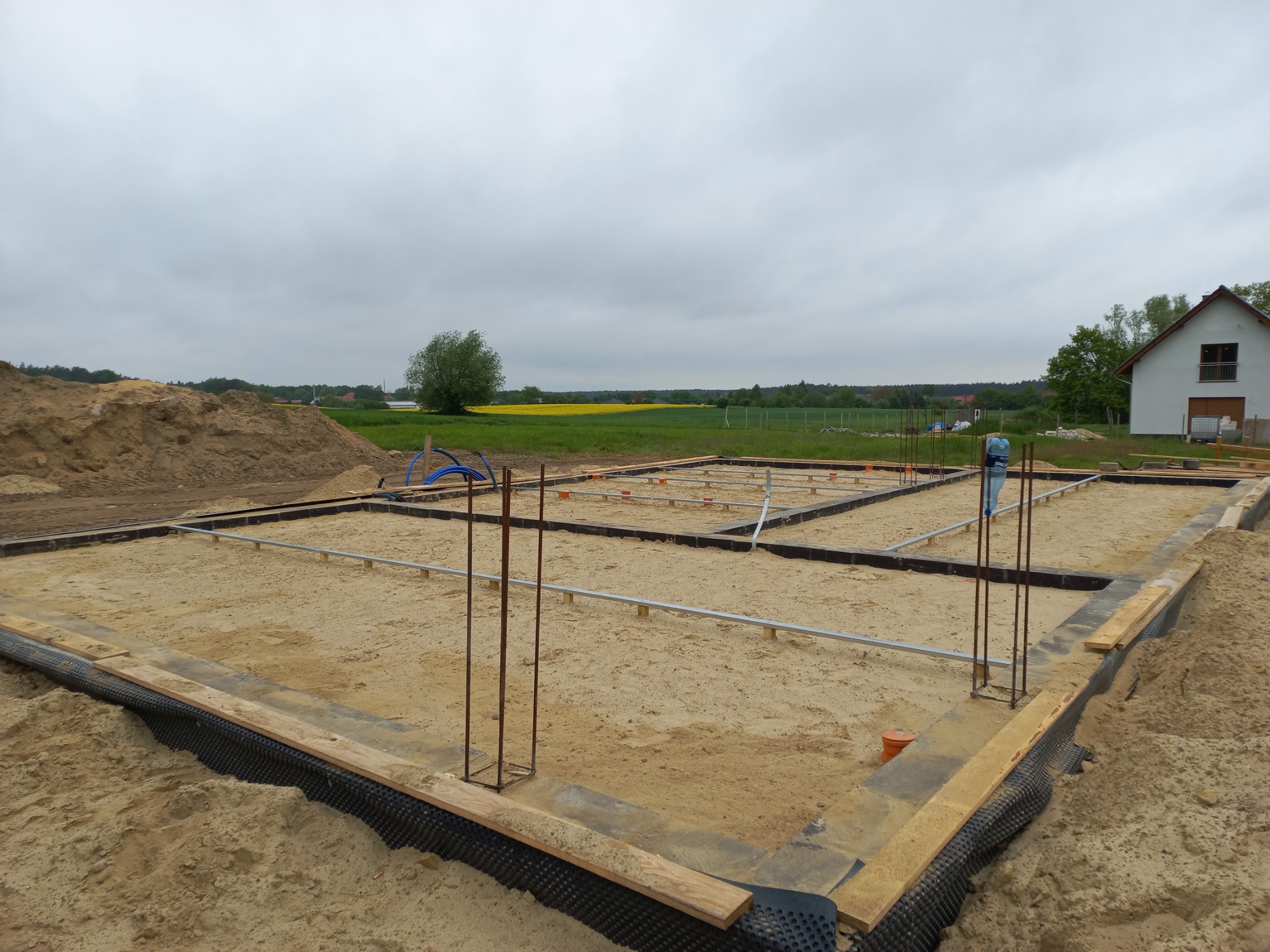 Fundamenty domu w trakcie budowy: szalunek z drewnianych desek, izolacja pionowa z folii kubełkowej, wystające pręty zbrojeniowe, widok na pole i dom w tle.