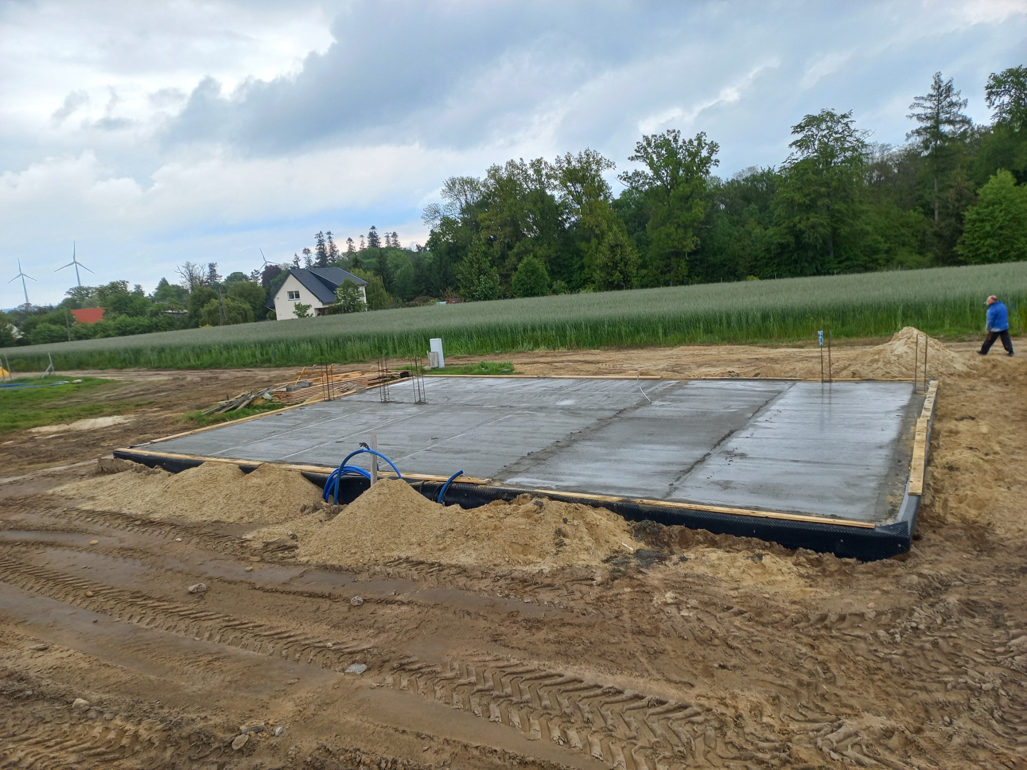 Wylana płyta fundamentowa pod budowę domu jednorodzinnego, widoczne wystające pręty zbrojeniowe, niebieski wąż do podlewania oraz hałdy piasku, w tle pola uprawne, dom i turbiny wiatrowe.