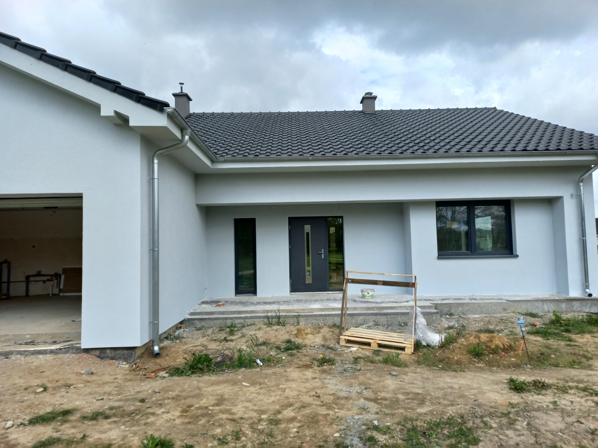 Nowoczesny dom parterowy z szarą elewacją i grafitową dachówką, widoczne elementy wykończeniowe, teren wokół domu w trakcie prac ziemnych.