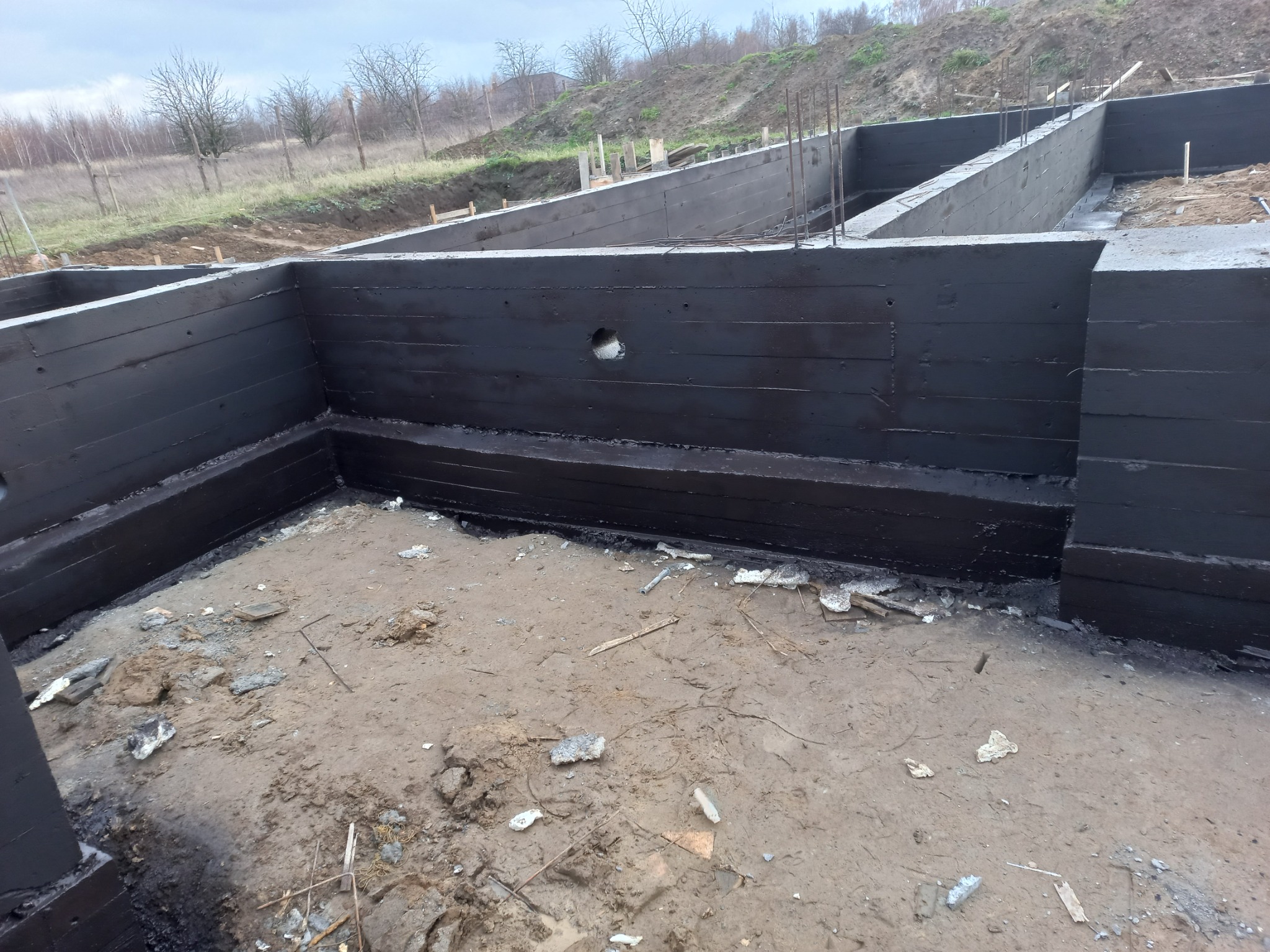 Monolityczny fundament zbrojony stalą, zabezpieczony hydroizolacją bitumiczną, widoczny otwór technologiczny w ścianie fundamentowej, teren budowy z resztkami materiałów.