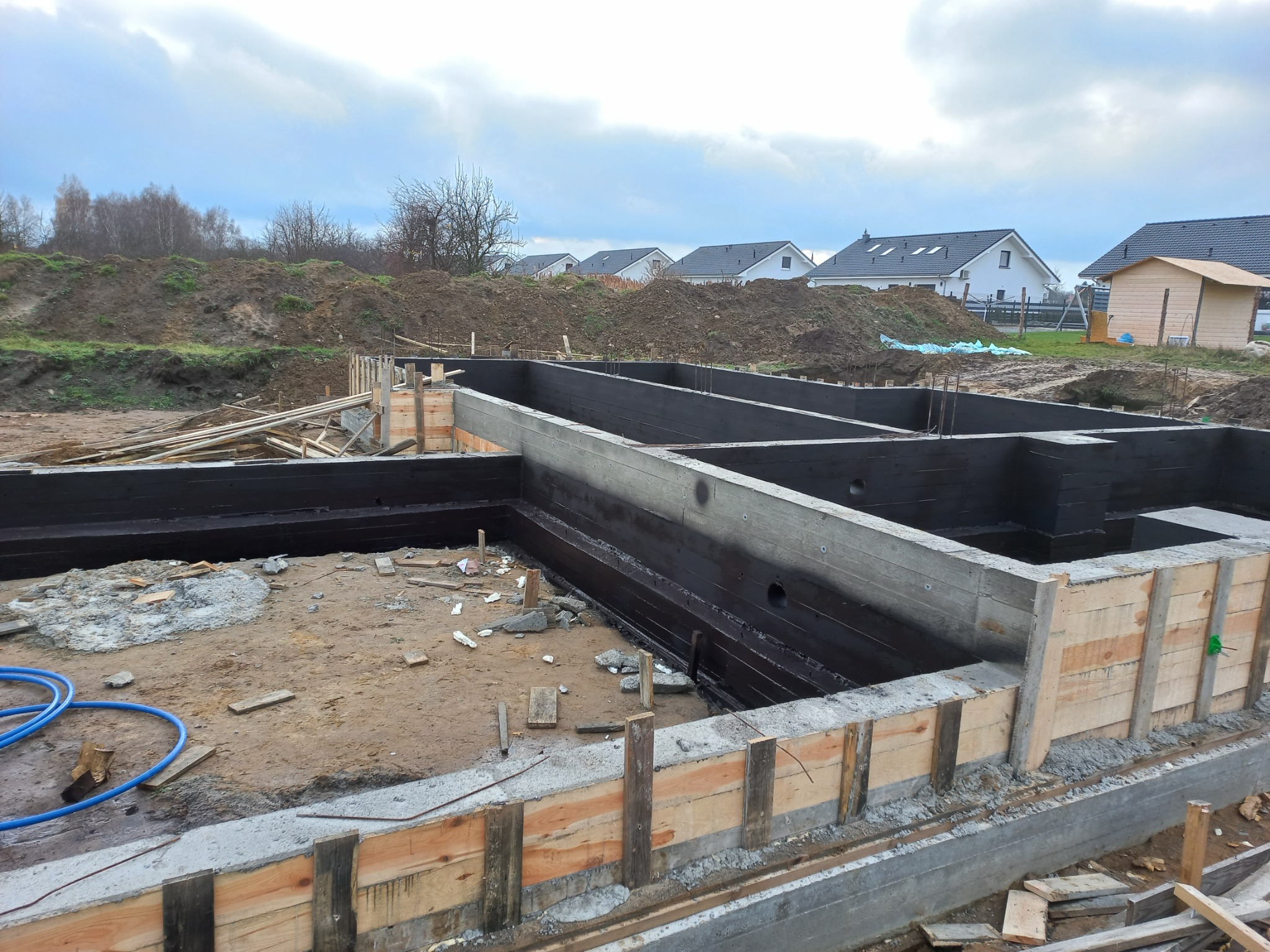 Monolityczny fundament z izolacją bitumiczną, szalunek z desek, widoczne zbrojenie i instalacja niebieskich rur, w tle ziemia i zabudowa jednorodzinna pod pochmurnym niebem.