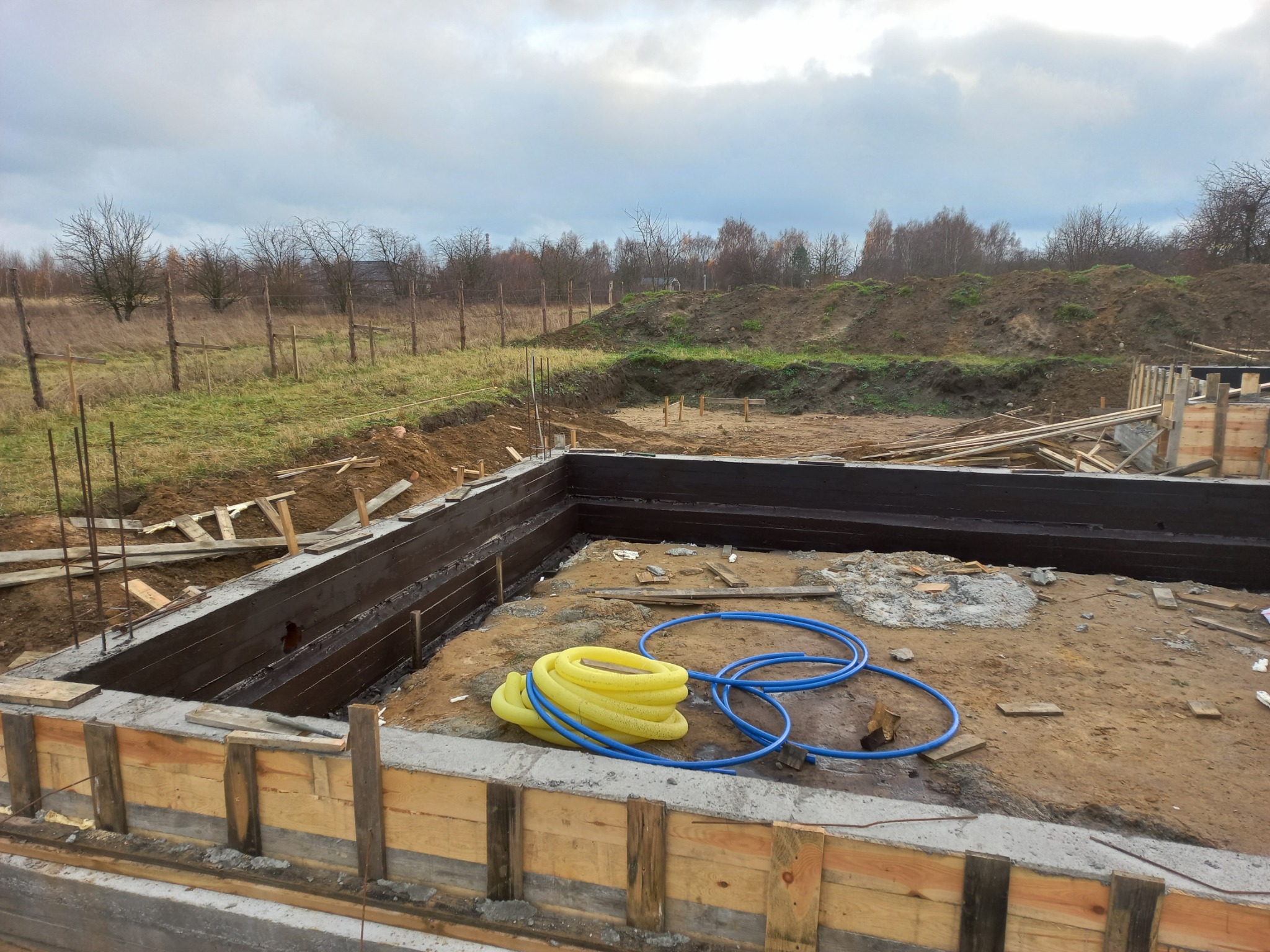 Fundament domu murowanego z hydroizolacją, szalunkiem i rurami instalacyjnymi na placu budowy w pochmurny dzień.
