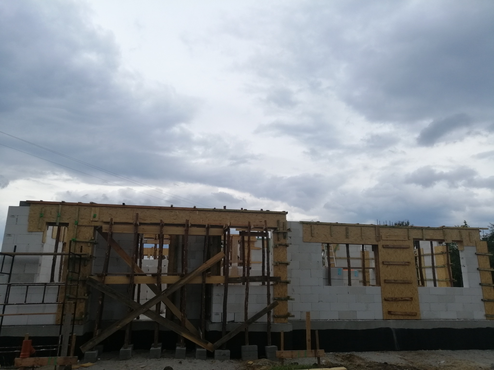 Budowa domu w stanie surowym otwartym z bloczków, widoczne drewniane szalunki okienne i podpory, pochmurne niebo w tle.