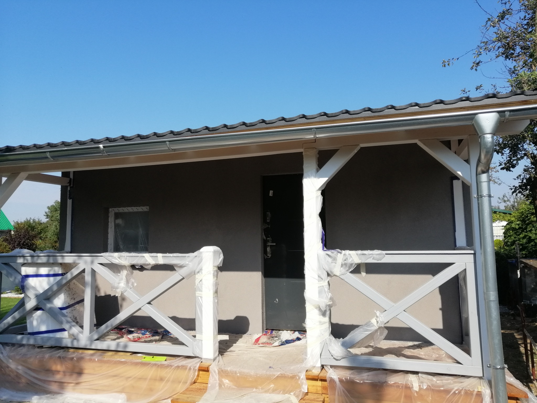 Altana ogrodowa z szarą elewacją i białą, drewnianą balustradą w trakcie prac wykończeniowych, zabezpieczona folią malarską. Widoczny fragment dachu z rynną i błękitne niebo.