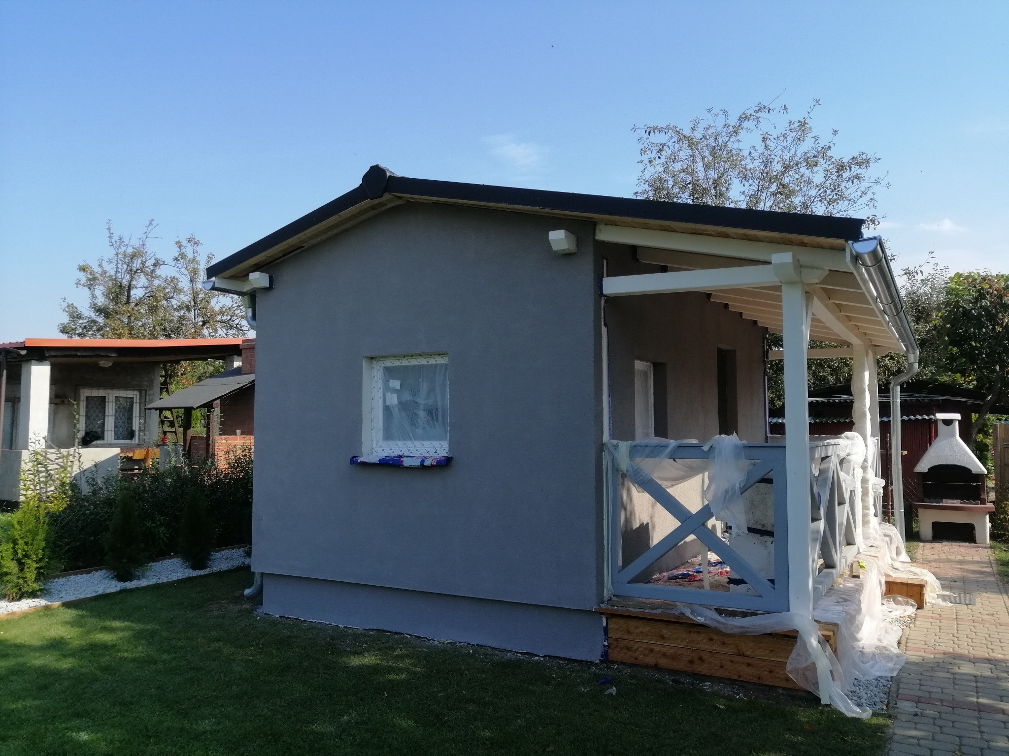 Szara altana ogrodowa z białymi elementami wykończeniowymi, częściowo zabezpieczona folią malarską podczas prac wykończeniowych elewacji i balustrady.