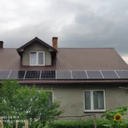 Dom jednorodzinny z zamontowanymi panelami słonecznymi na brązowym, metalowym dachu, widoczne drzewa i słonecznik w ogrodzie, pochmurne niebo.