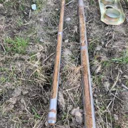 Dwa rdzenie glebowe w metalowych rurach, wyjęte z ziemi, leżą na trawie. Widoczne fragmenty gleby wewnątrz rur i metalowy uchwyt obok. Próbki gleby do analizy.