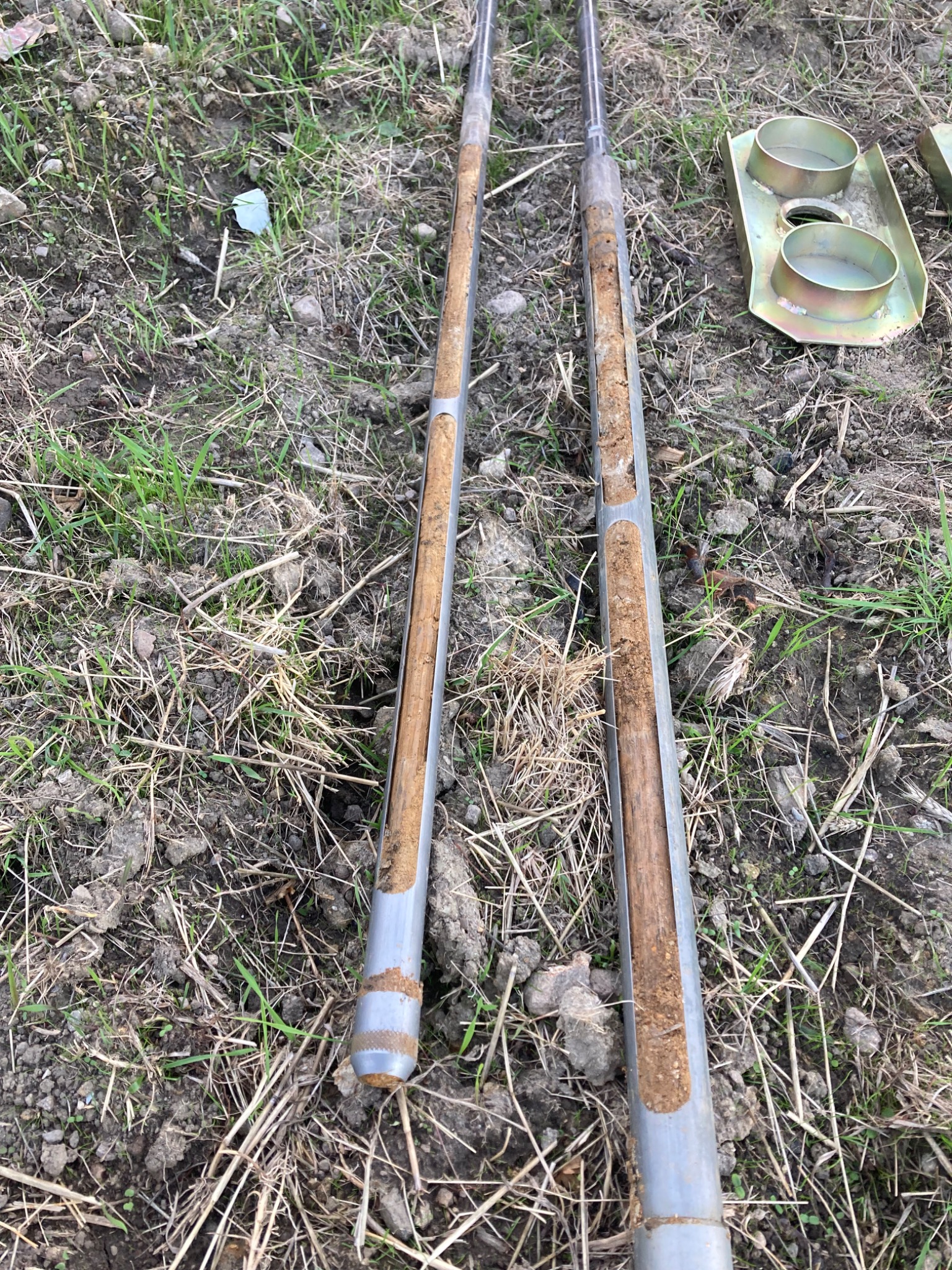 Dwa rdzenie glebowe w metalowych rurach, wyjęte z ziemi, leżą na trawie. Widoczne fragmenty gleby wewnątrz rur i metalowy uchwyt obok. Próbki gleby do analizy.